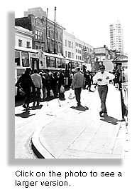 People are getting onto the buses on Stockton Street. They waited for twenty minutes for a bus to come. Here comes two buses fighting through the traffic of Chinatown. People of all ages have crowed around the doors trying to get on.