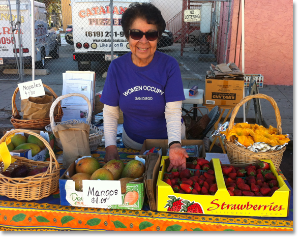 Virginia Franco showing off fresh fruits and squash blossoms (on the right) at the Women Occupy San Diego (WOSD) booth at Sobreruedas (30th and Imperial) in San Diego. Photo by Nic Paget-Clarke.'
